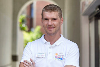 Hörgeräteakustiker in Arbeitskleidung steht mit verschränkten Armen in einem Gang