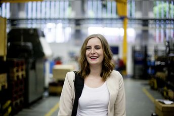 Junge Frau steht im einer Kameratasche über der Schulter in einer Fabrikhalle