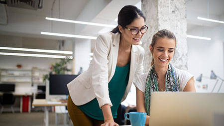 Zwei junge Frauen, ein stehend, eine sitzend, schauen auf einen Computerbildschirm